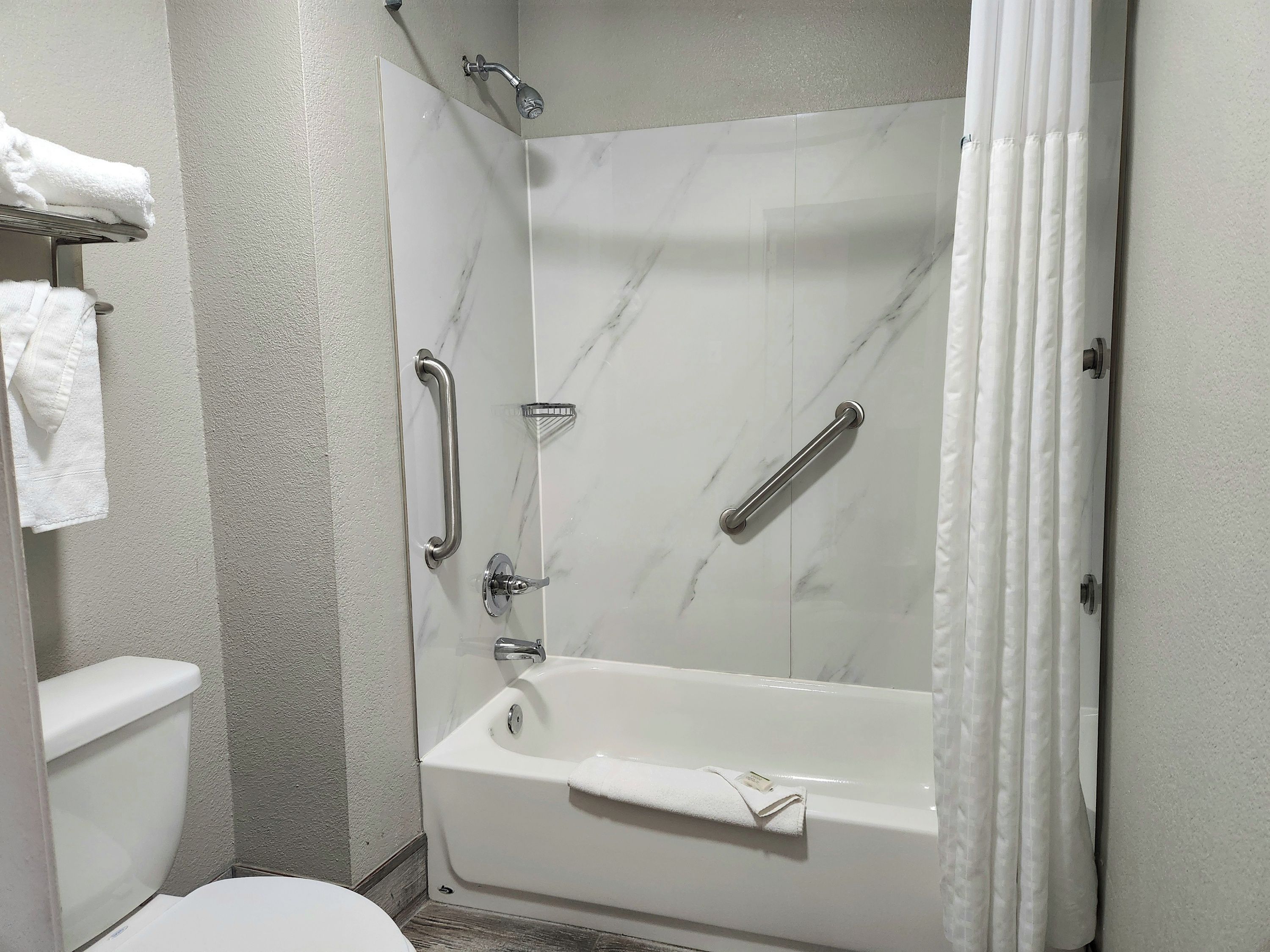 A bathroom with a white bathtub, a shower with a handheld showerhead, a grab bar, and a toilet.
