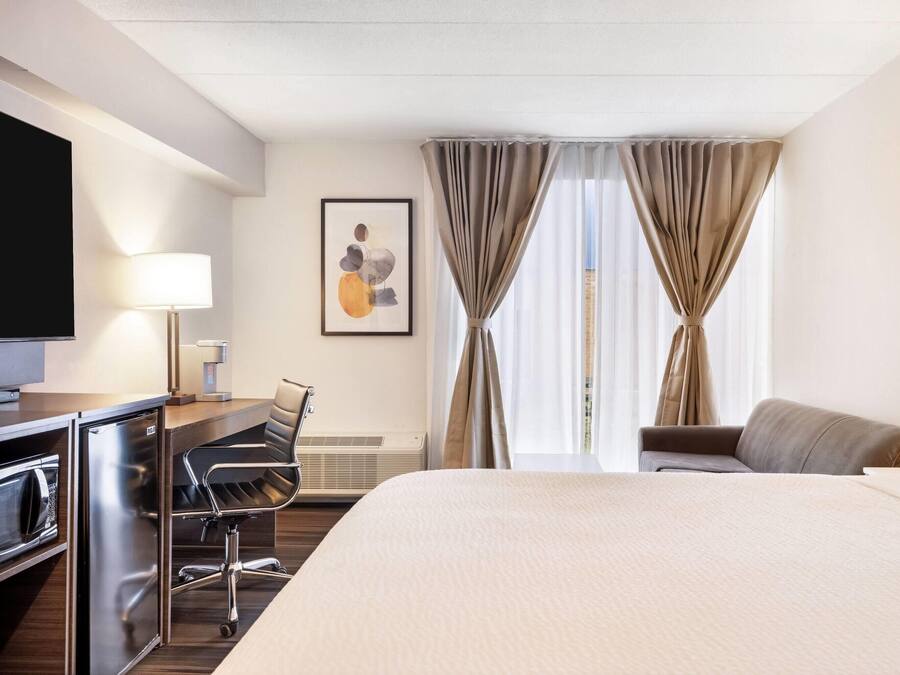 A hotel room with a bed, a desk with a computer, a chair, a TV, and a window with curtains.