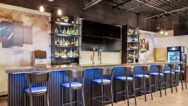 Zona de bar con mostrador, varios taburetes, estantes con botellas y un refrigerador.