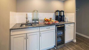 Una cocina con gabinetes blancos, encimera de granito, pileta, cafetera, frutero y refrigerador.