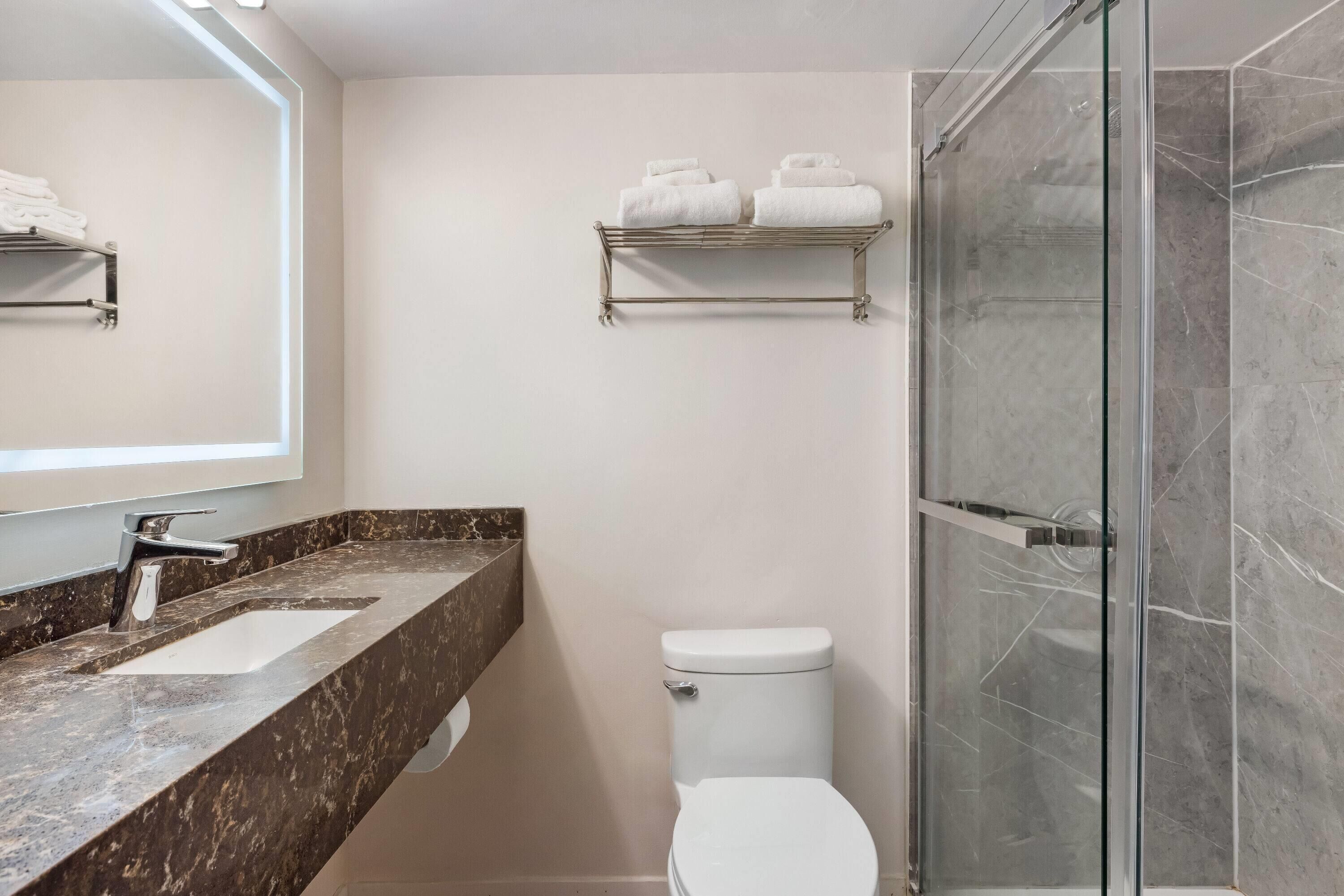 A bathroom with a marble countertop, a white sink, a toilet, a glass shower enclosure, and a towel rack with towels.