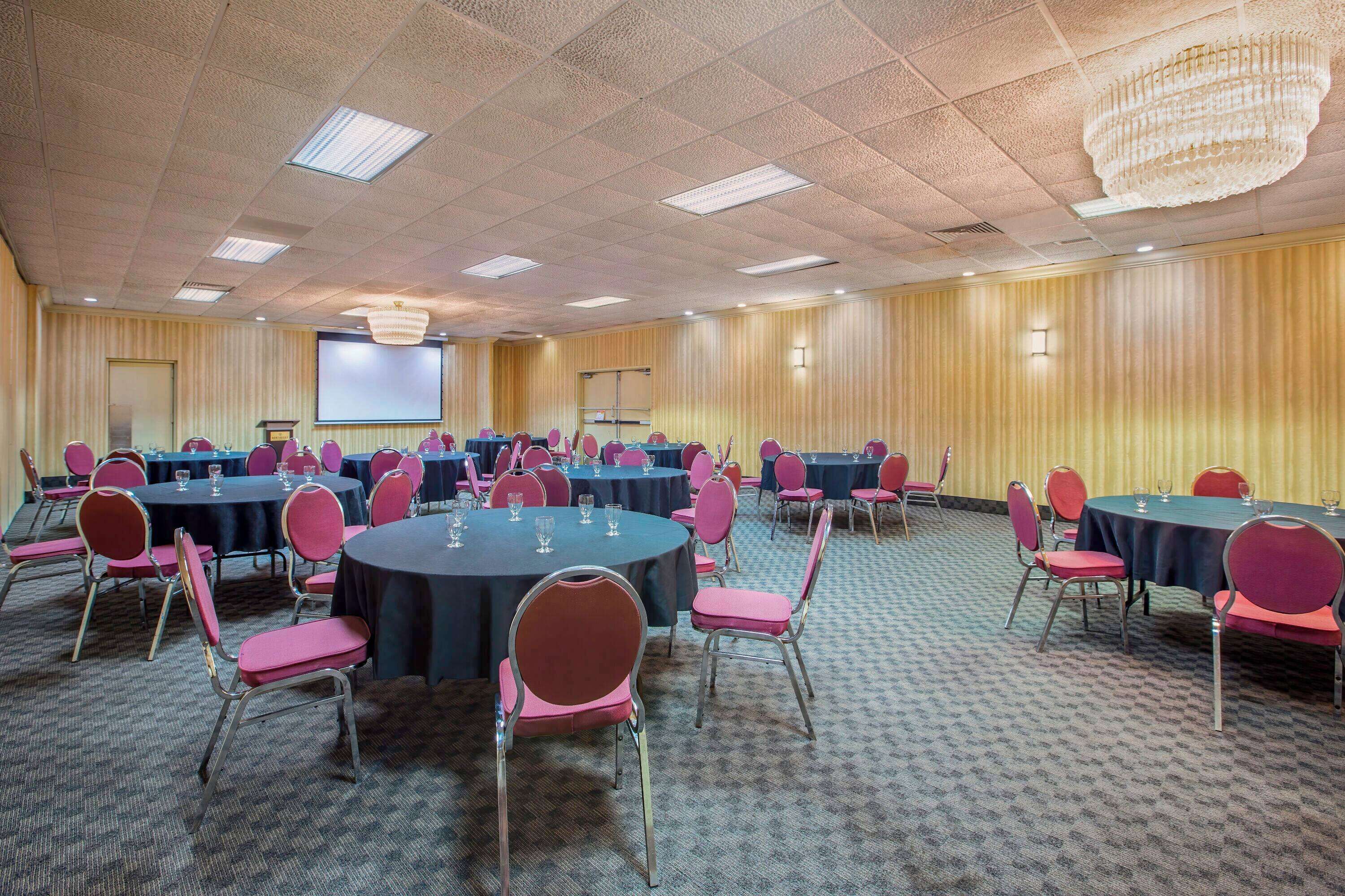 Une salle de conférence équipée de tables et de chaises rondes, d’un écran de projection et d’un lustre.