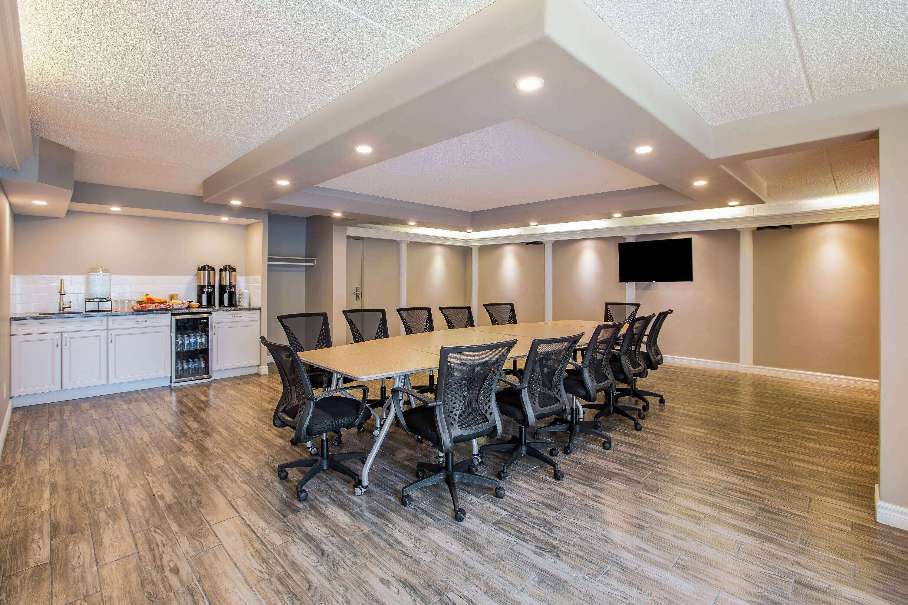 A conference room with a long table, chairs, a kitchen area with cabinets and appliances, and a flat-screen TV on the wall.