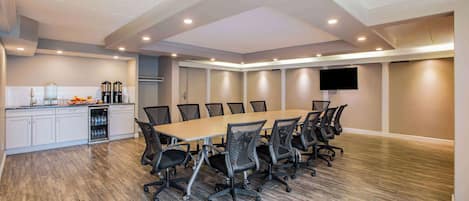A conference room with a long table, chairs, a kitchen area with cabinets and appliances, and a flat-screen TV on the wall.