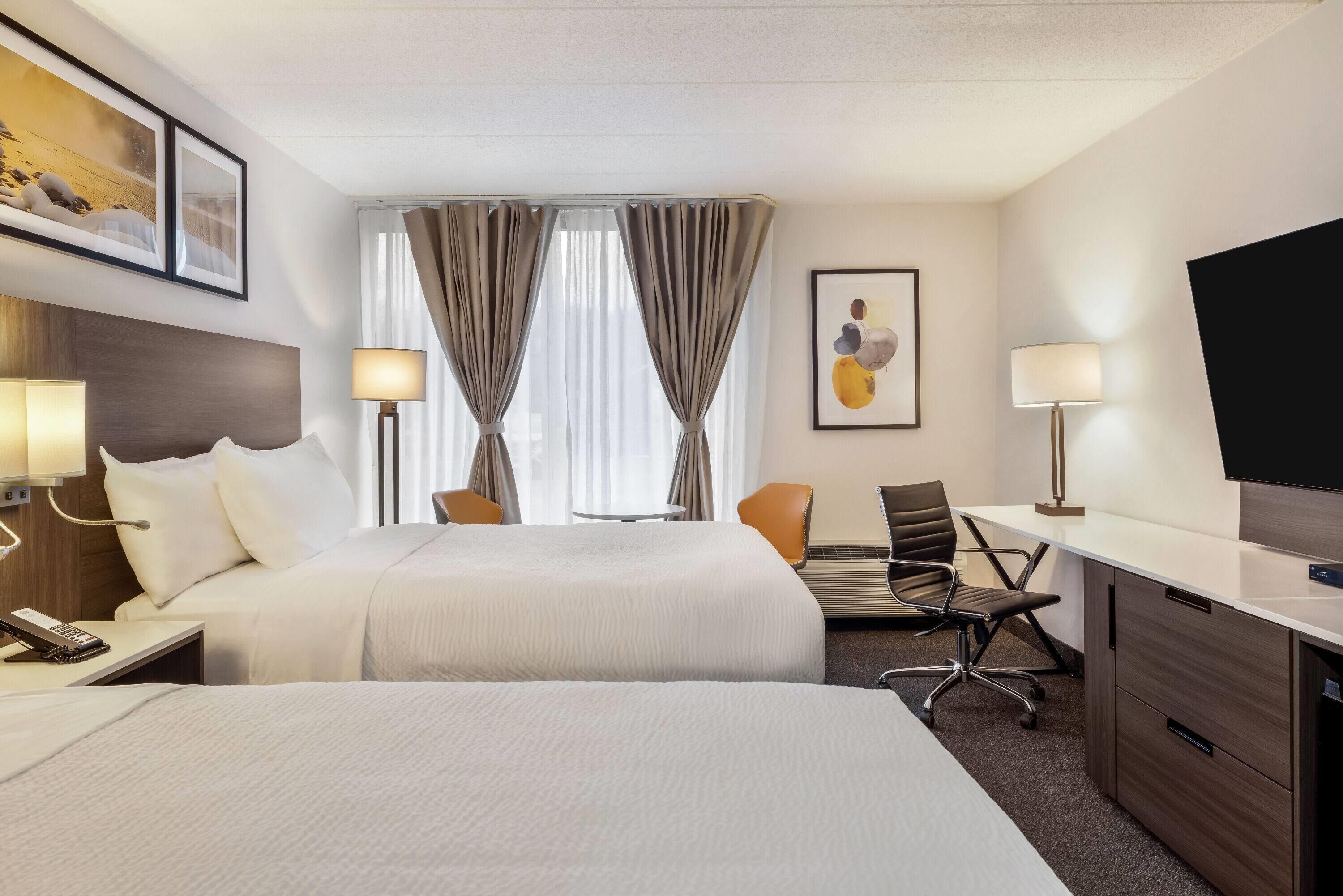 A hotel room with two beds, a desk, a chair, a TV, and a lamp.