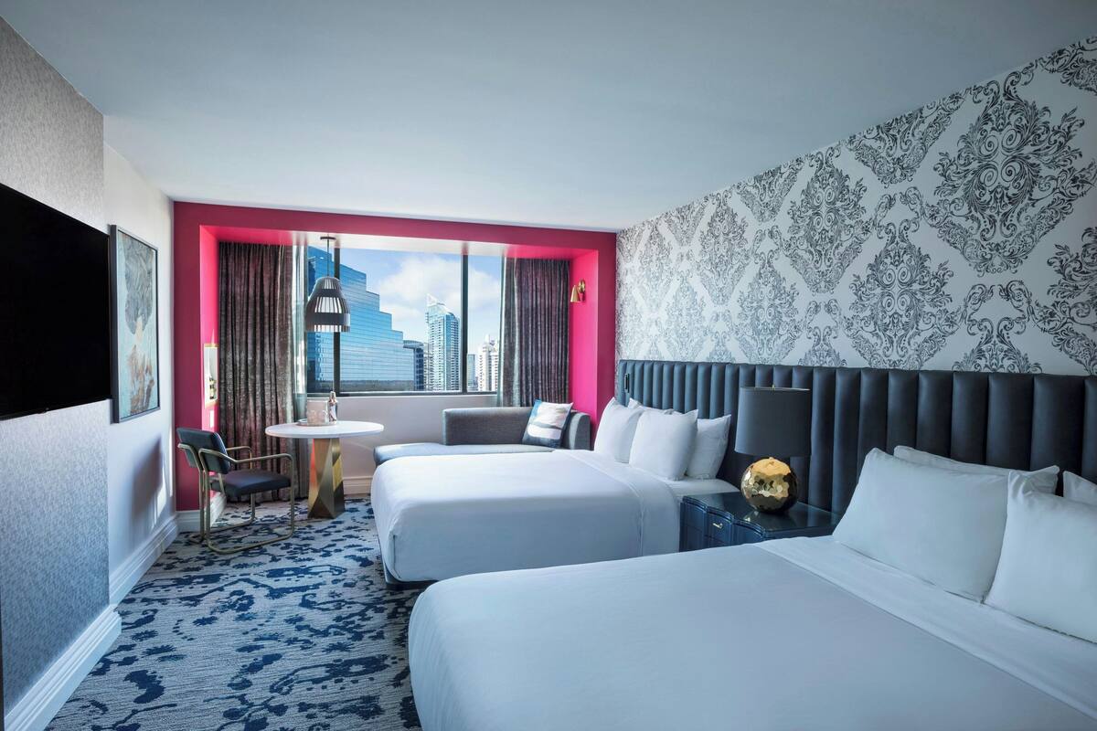 A hotel room with two beds, a small table, a chair, and a view of the city skyline.