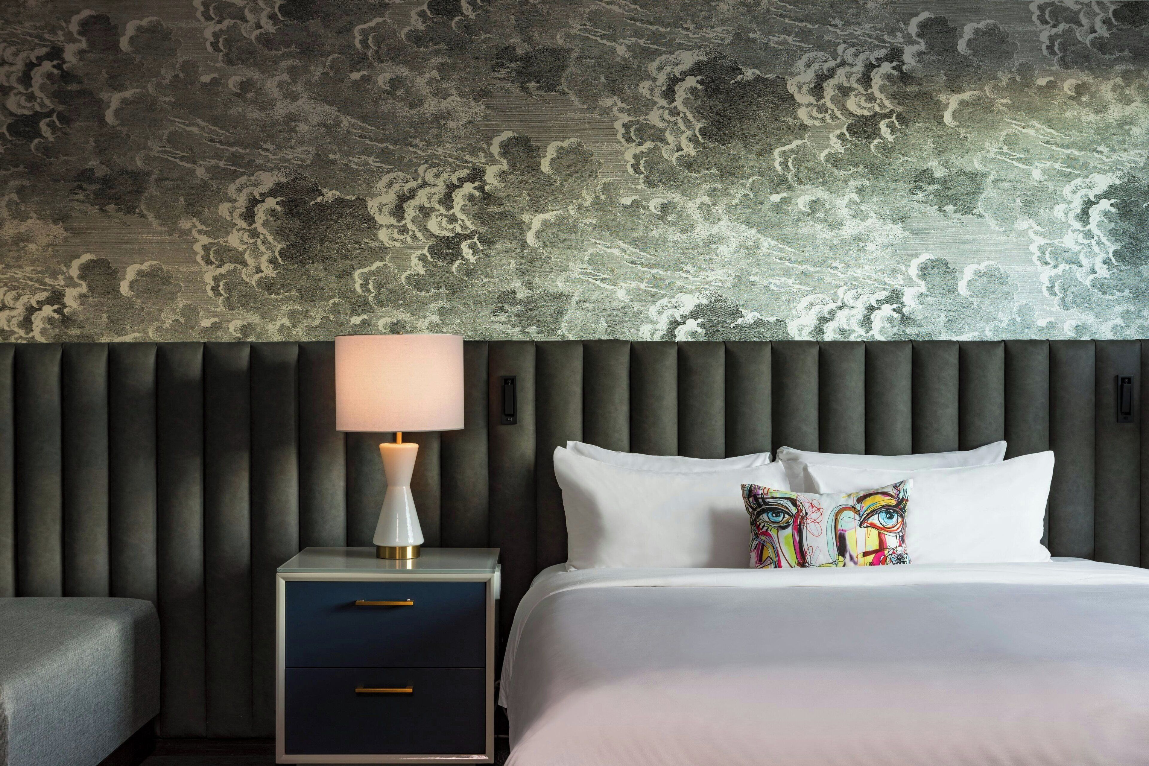 A hotel room with a cloud-patterned ceiling, a modern bed with white linens, a bedside table with a lamp, and a dark-colored nightstand with drawers.