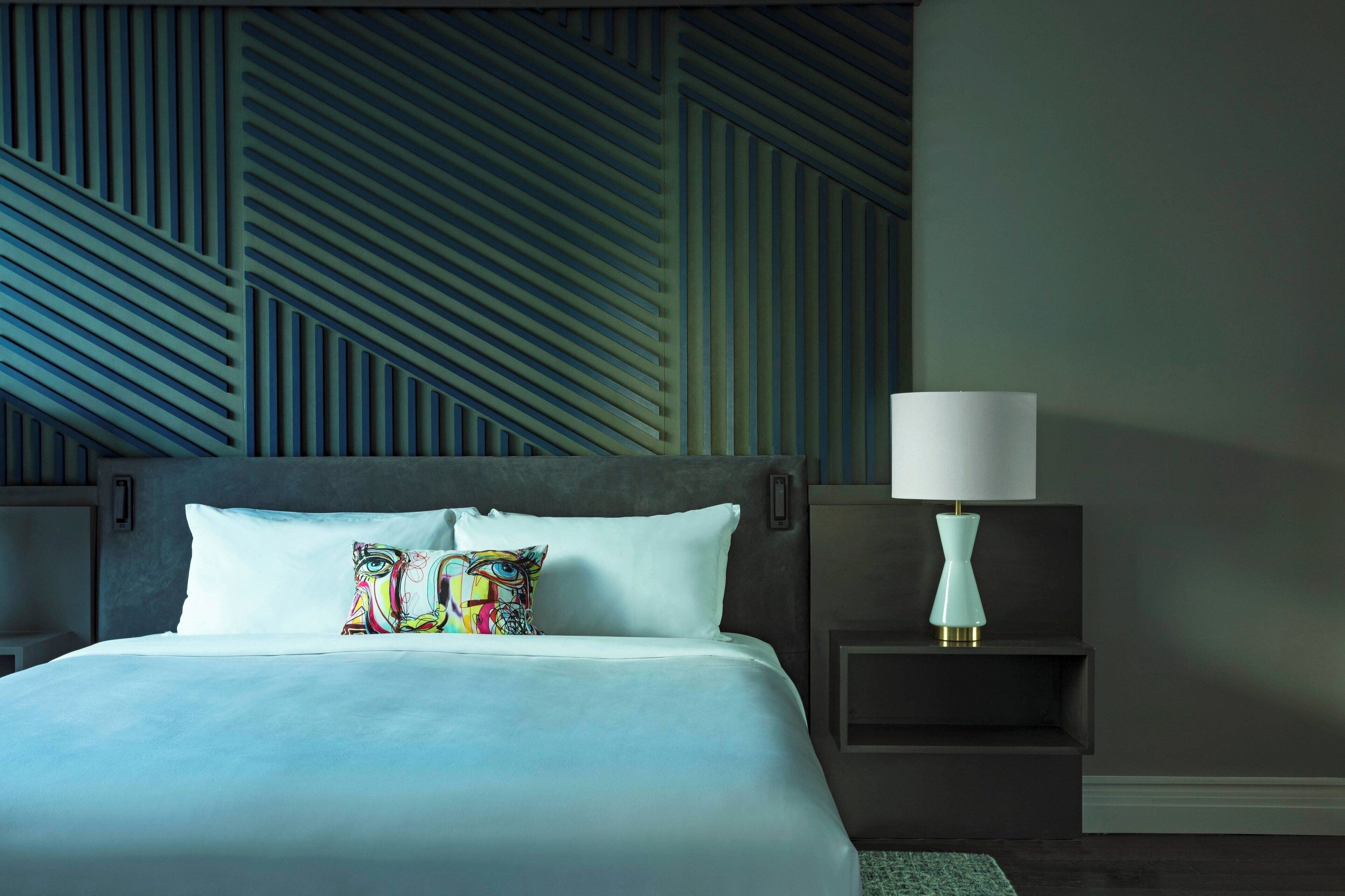 Una habitación de hotel moderna con una cama grande, un cojín colorido, una mesita de noche con una lámpara y un papel tapiz de diseño geométrico.