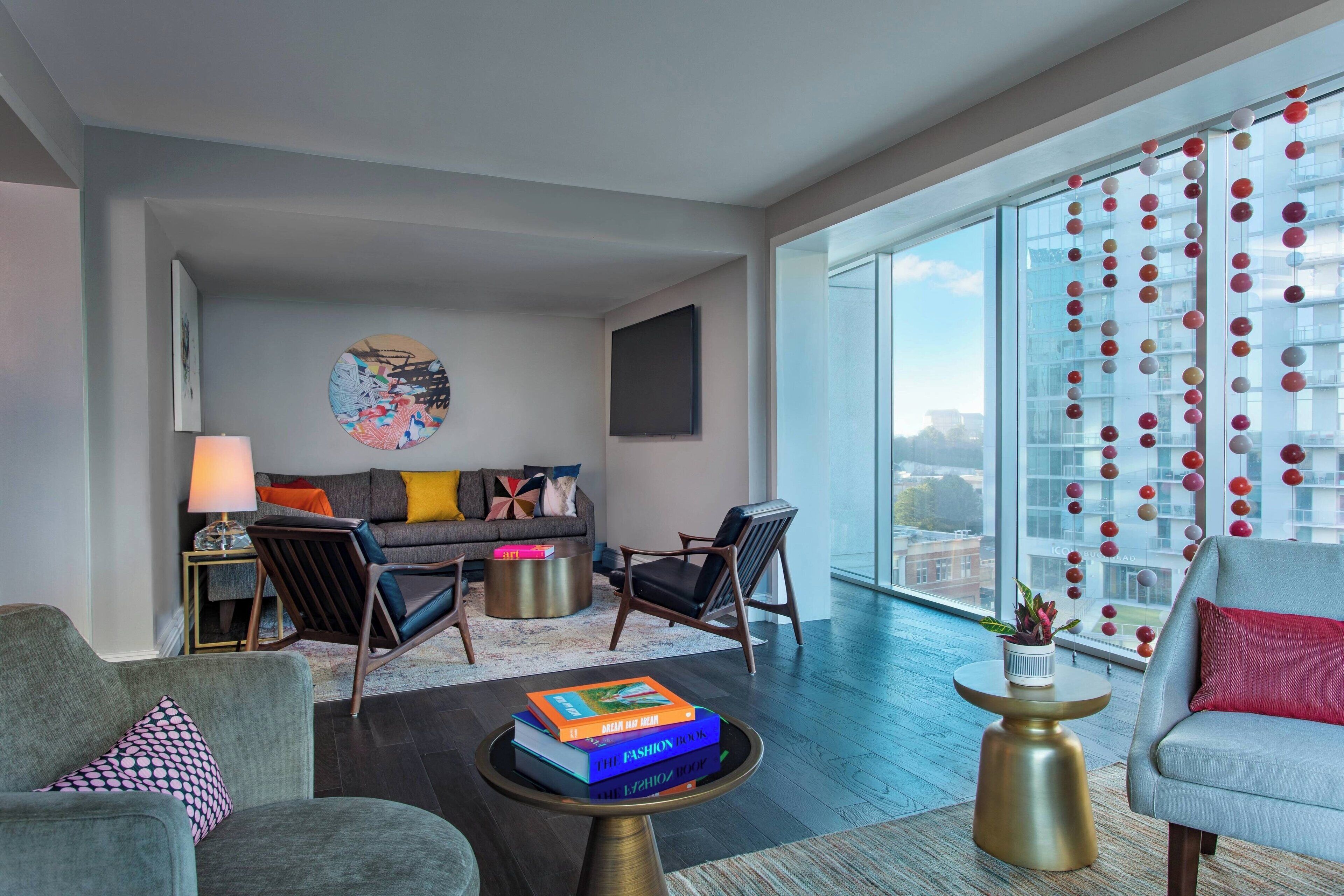 A modern living room with a sofa, chairs, a coffee table with books, a lamp, and a large window with a view of the city.