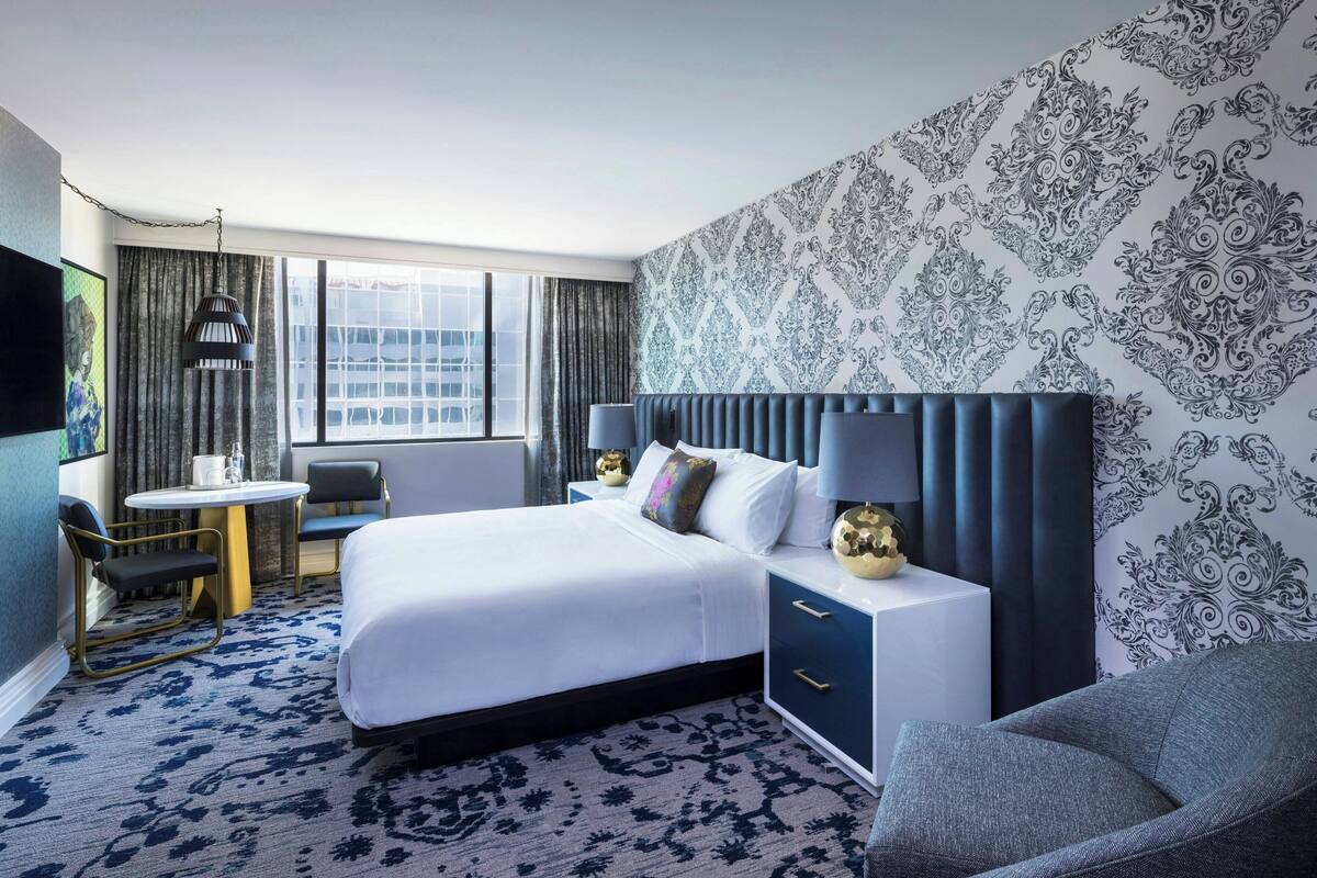 A hotel room with a large bed, a desk, and a chair. There is a window with curtains, a wall with a patterned wallpaper, and a television on the wall.