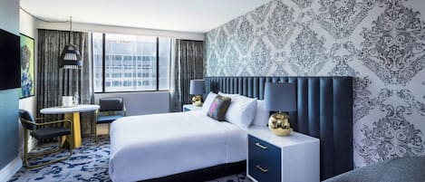 Habitación de hotel con una cama grande, un escritorio y una silla. Se ve una ventana con cortinas, una pared con papel tapiz estampado y un televisor en la pared.