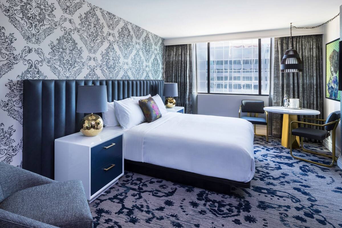 A hotel room with a large bed, a bedside table with lamps, a chair, a small table with a chair, and a view of the city through the window.