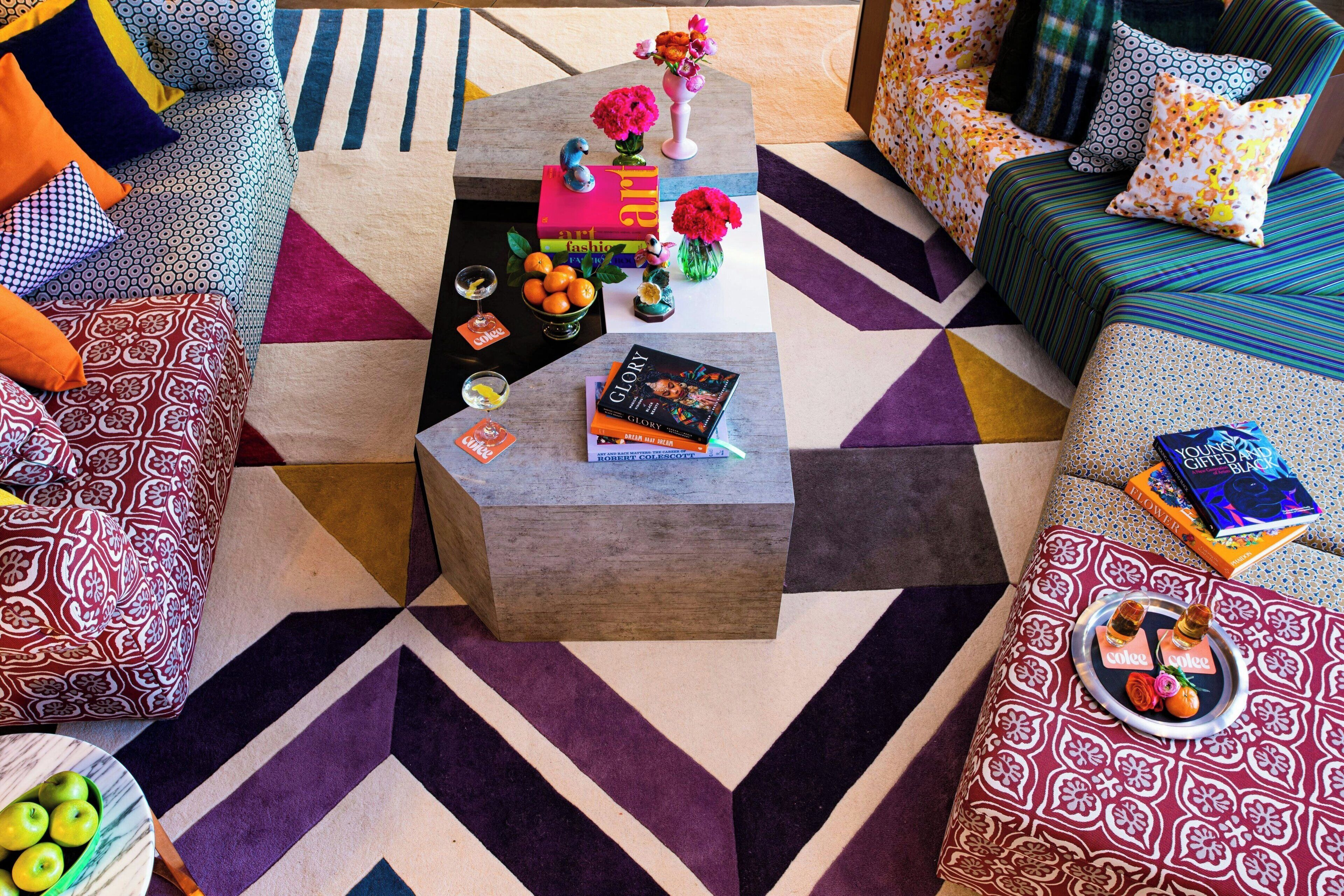Una sala de estar moderna con un tapete de diseño geométrico, una mesa de centro con libros y objetos decorativos, y un sofá con cojines estampados.