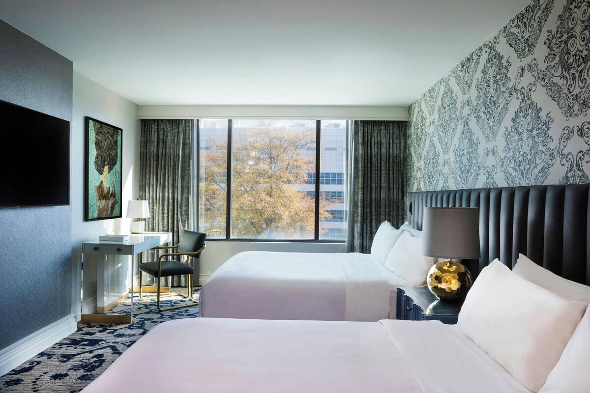A hotel room with two beds, a desk, a chair, a lamp, and a view of trees through the window.