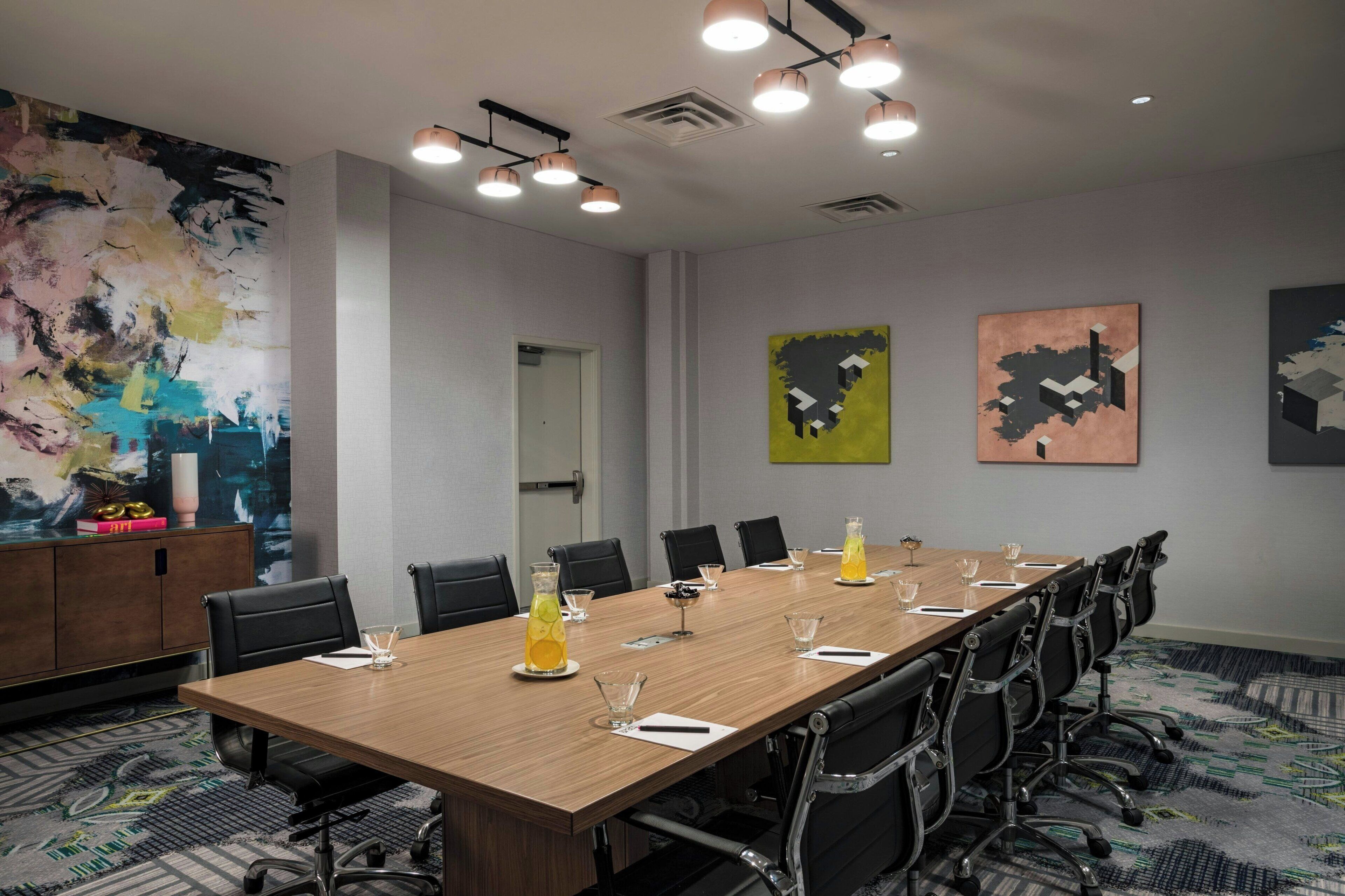 Un salón de conferencias con una larga mesa, sillas negras y una pared decorativa con arte abstracto.