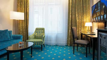 A hotel room with a blue carpet, a sofa, a striped armchair, a wooden chair, a small table, and a TV screen displaying a welcome message.