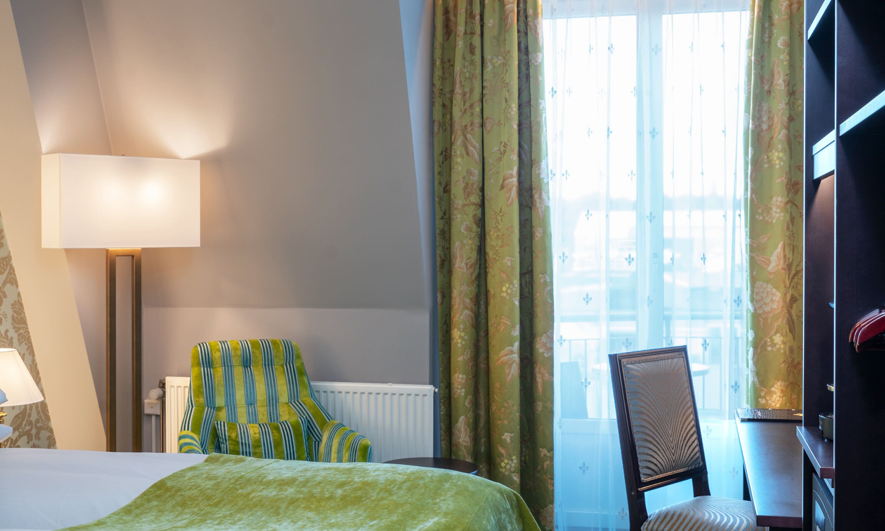 Una habitació d'hotel amb llit, cadira, taula, làmpada i finestra amb cortines.