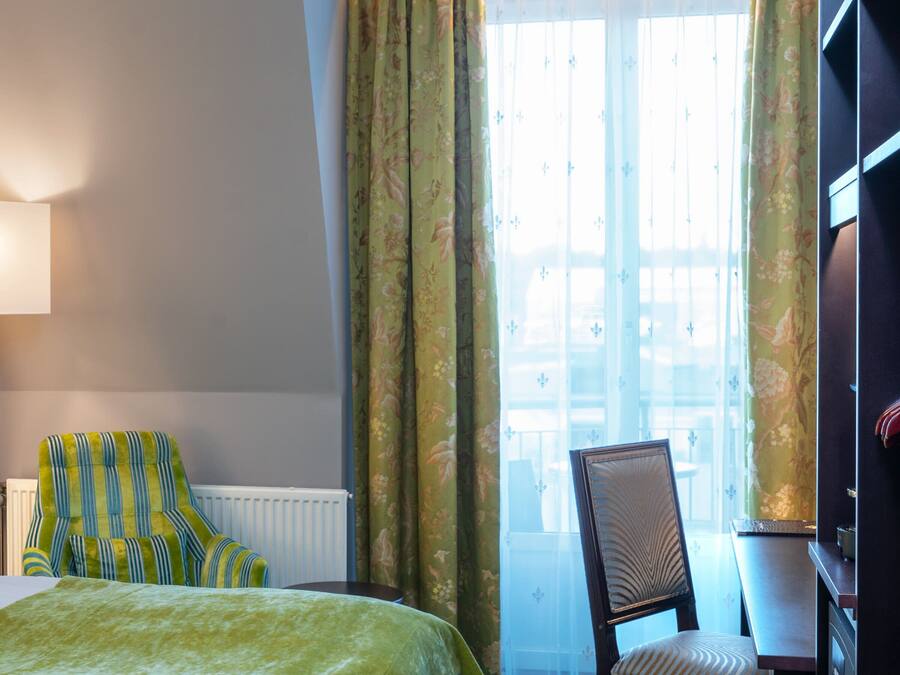 Una habitació d'hotel amb llit, cadira, taula, làmpada i finestra amb cortines.