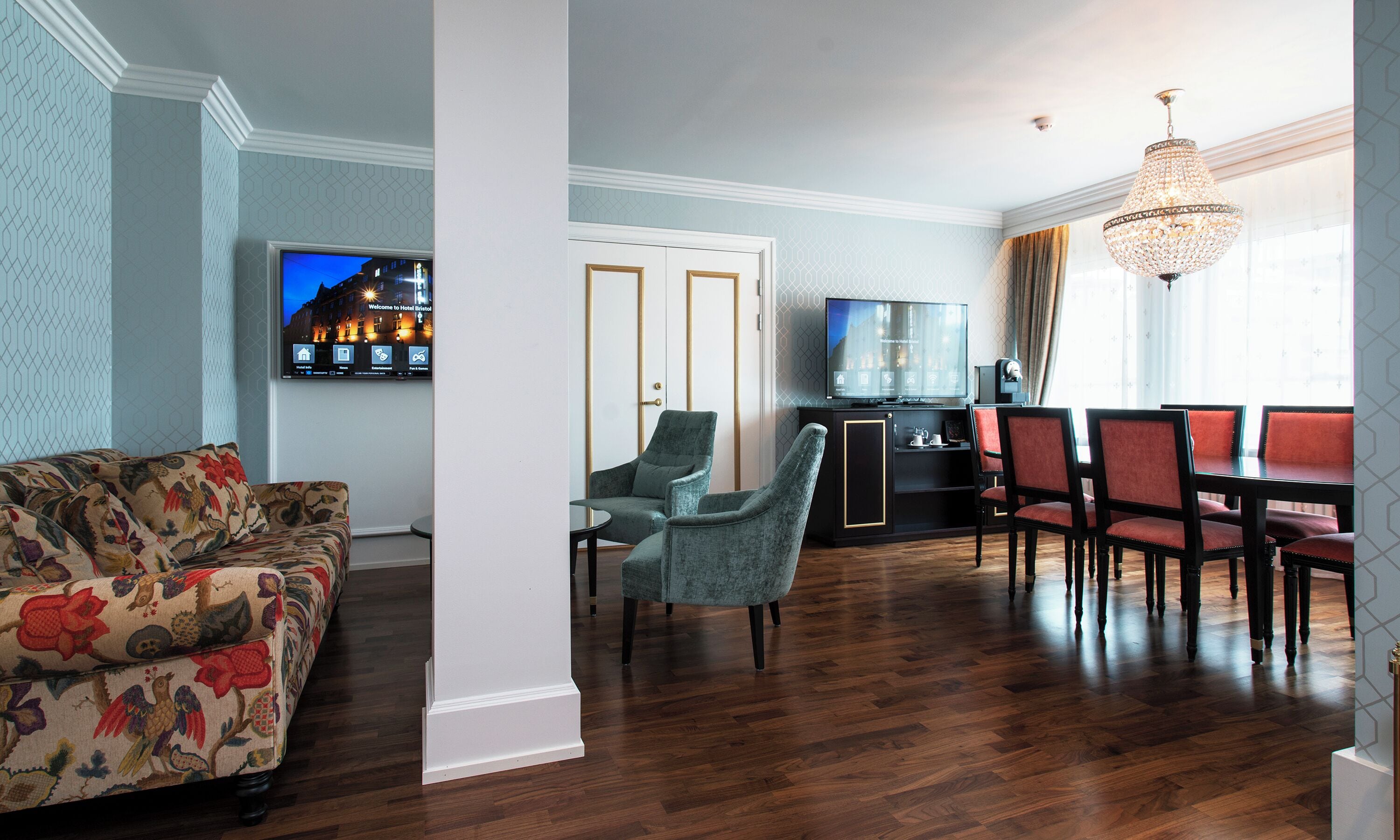 Habitación de hotel con piso de madera, sofá estampado con motivos florales, televisor, candelabro y zona de comedor con sillas.