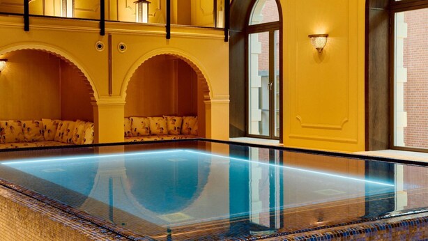 A large indoor swimming pool with a tiled edge, arched seating areas, and wall-mounted lighting. - Hotel Bristol (Oslo)
