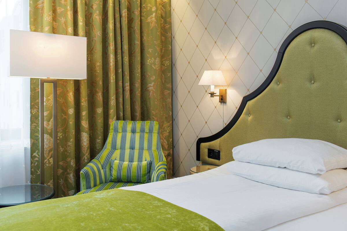 A hotel room with a bed, a striped armchair, a small table, a lamp, and curtains with a floral pattern.