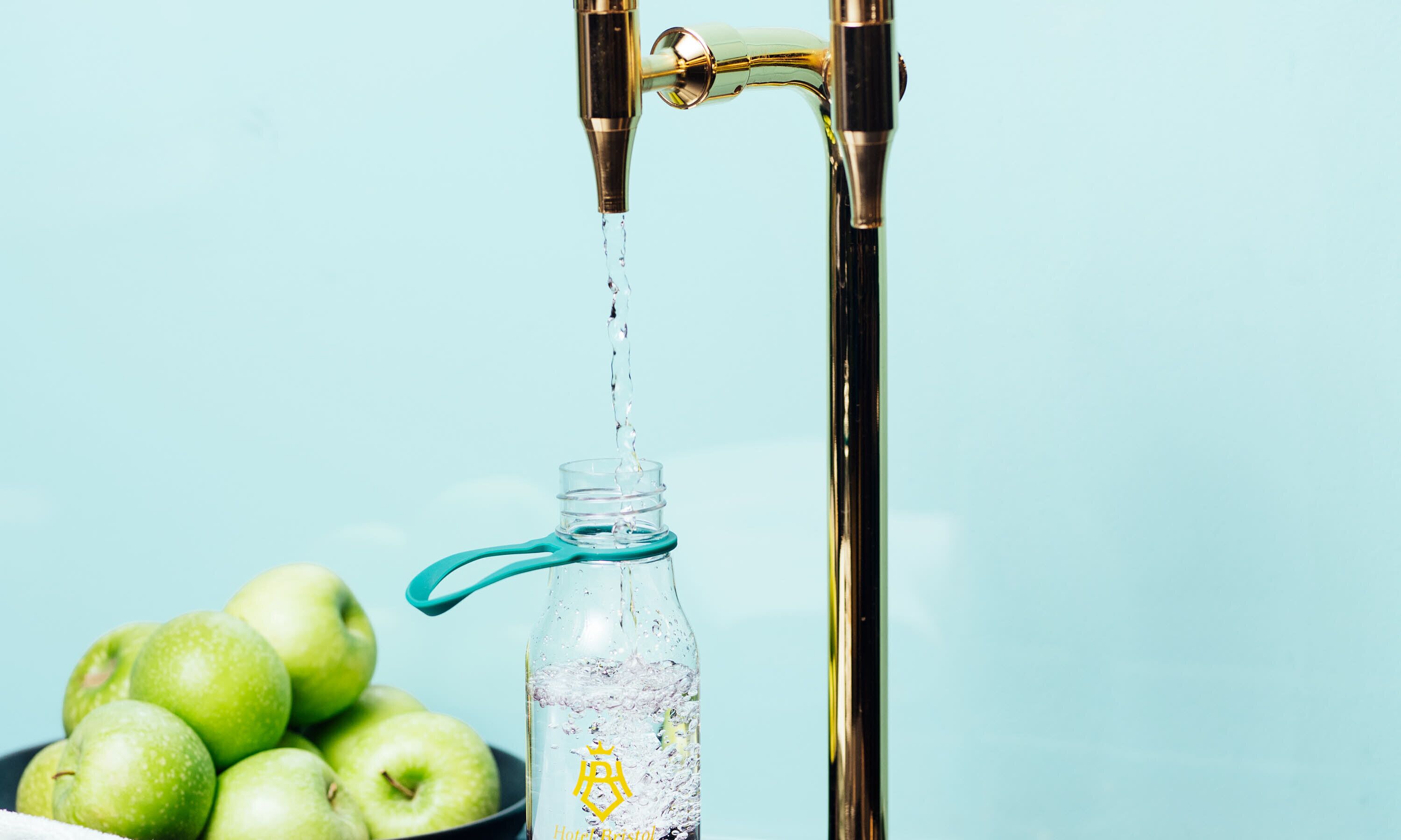 Een glazen fles met een blauwe hendel onder een stromende kraan, met groene appels op de achtergrond.