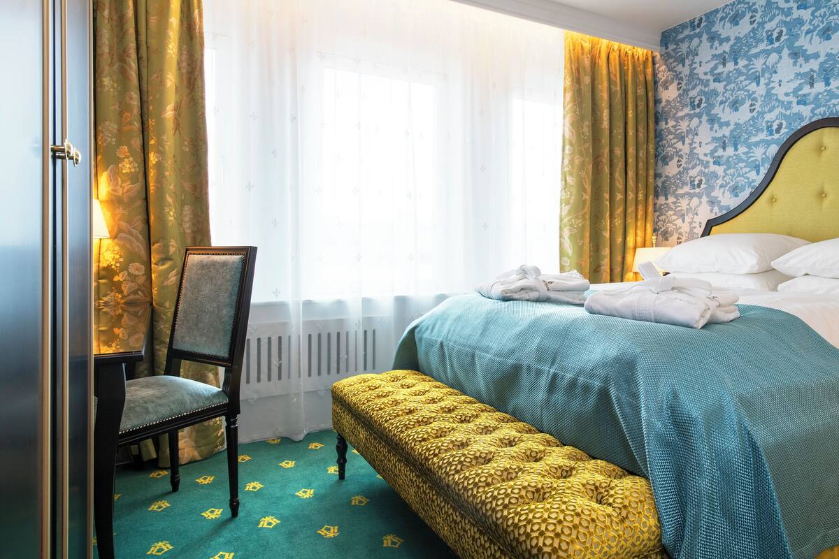 A hotel room with a bed, a chair, a bench, and a window with curtains.