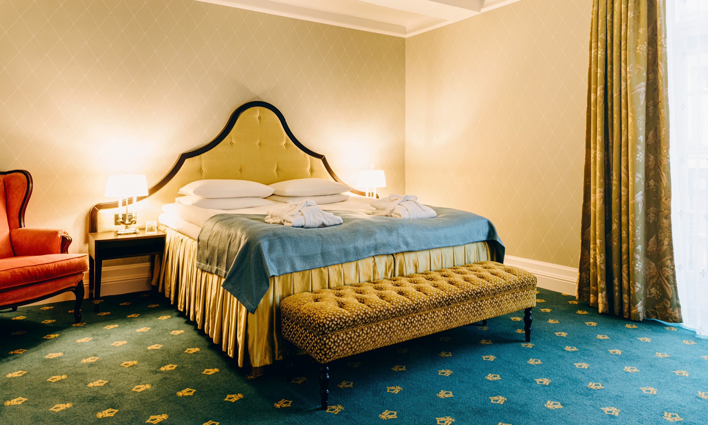 A hotel room with a large bed, a bench, a chair, and a nightstand with a lamp.