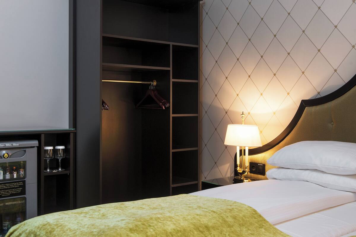 A hotel room with a bed, a nightstand with a lamp, a wardrobe, and a minibar.