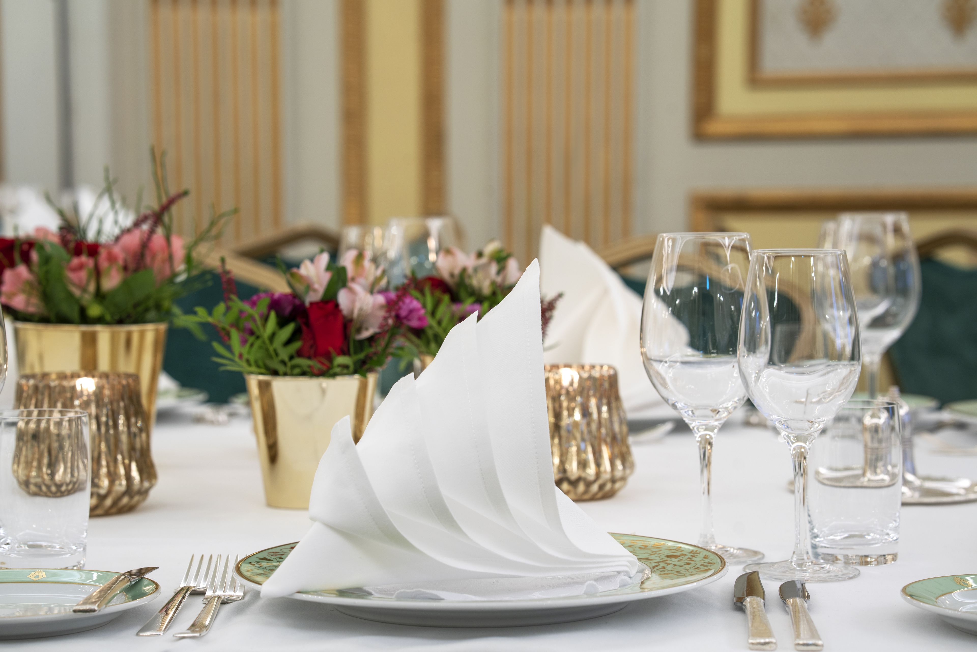 Ein Tisch ist mit weißen Servietten, klaren Weingläsern und goldenen Vasen mit Blumen gedeckt.