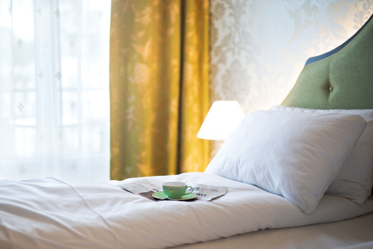 Green teacup on white bed linens with gold curtains behind