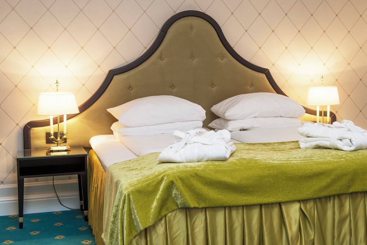 A neatly made bed with white linens and a green bedspread, two bedside lamps, and a nightstand with a lamp.
