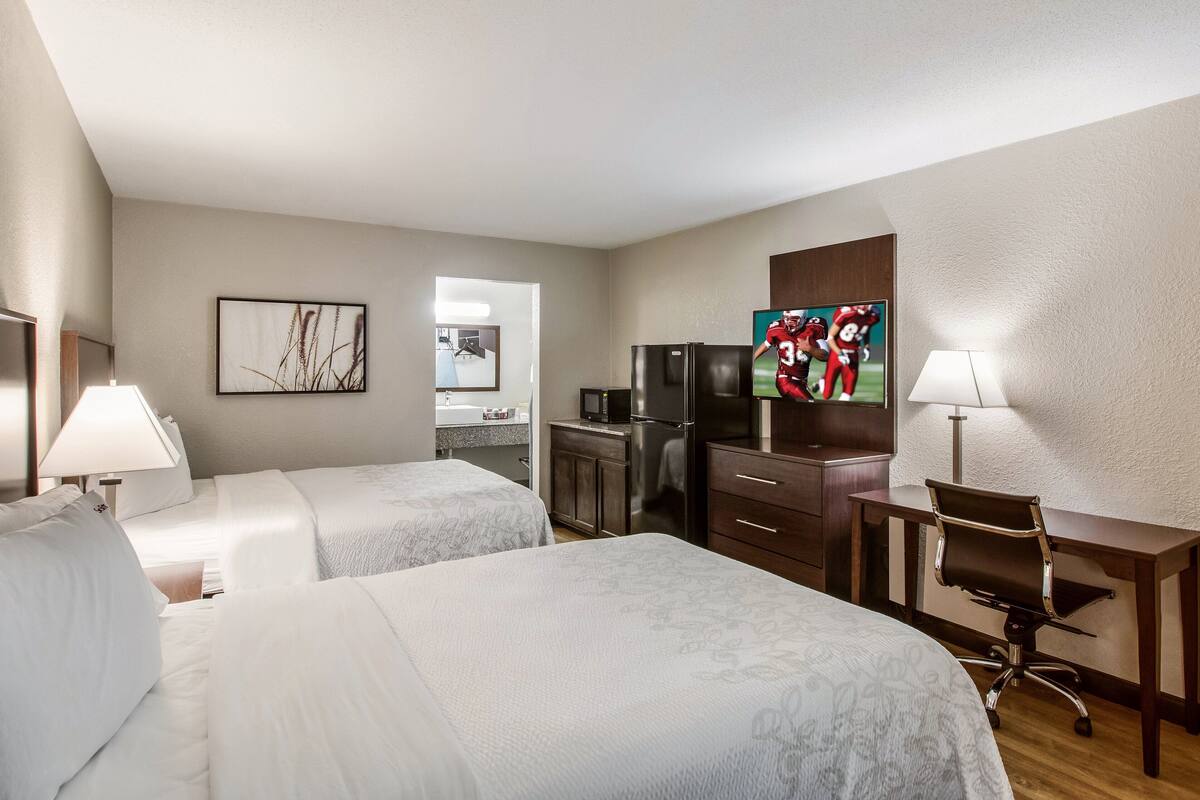 A hotel room with two beds, a desk, a refrigerator, a TV, and a bathroom visible in the background.