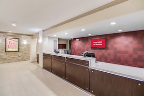 A hotel reception area with a reception desk, a digital screen displaying 'Free Wi-Fi All Rooms,' and a 'Red Roof Plus+' sign on the wall. - Red Roof Inn PLUS+ Dallas – Addison (Addison)