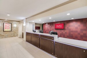Uma área de recepção de hotel com um balcão, uma tela digital exibindo 'Wi-Fi Gratuito em Todos os Quartos' e um letreiro 'Red Roof Plus+' na parede.