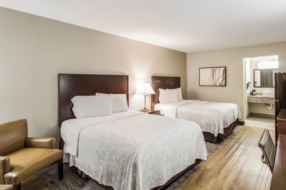 A hotel room with two beds, a chair, a desk, and a bathroom visible through an open door.