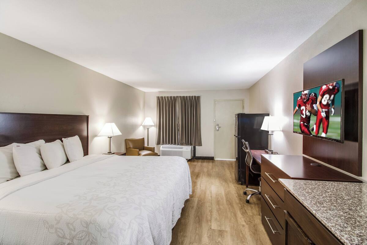 A hotel room with a large bed, a desk with a flat-screen TV, a chair, a lamp, and a window with curtains.