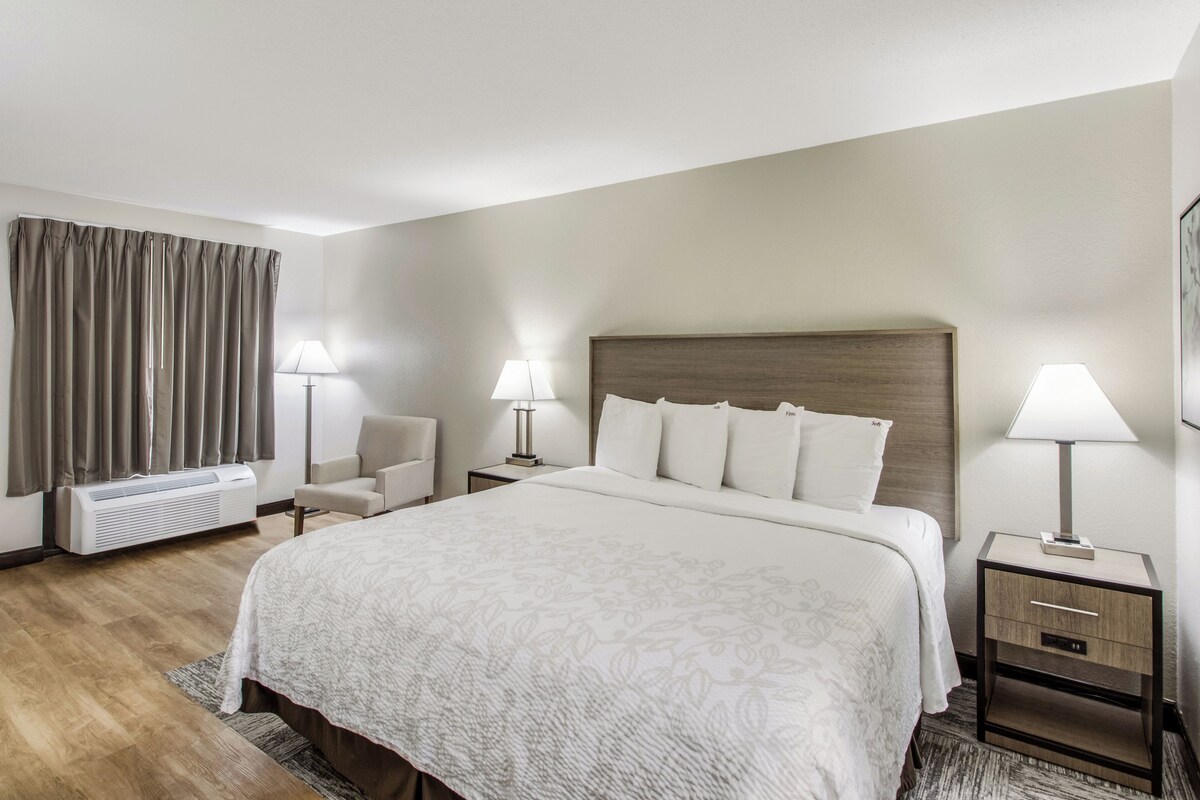 A hotel room with a large bed, two bedside tables with lamps, a chair, a desk, a desk lamp, and a window with curtains.
