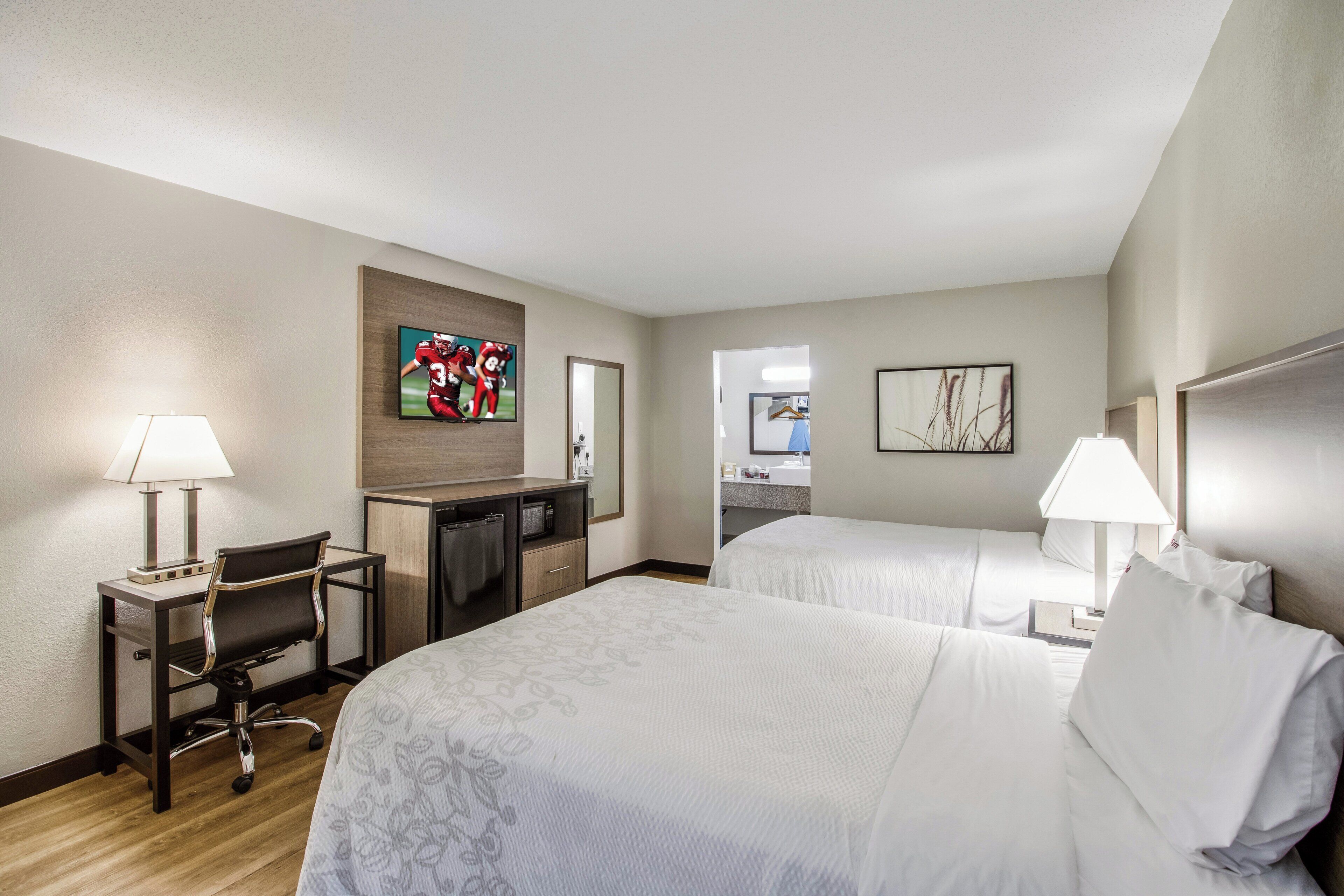 A hotel room with two beds, a desk with a chair, a TV, and a bathroom visible through an open door.