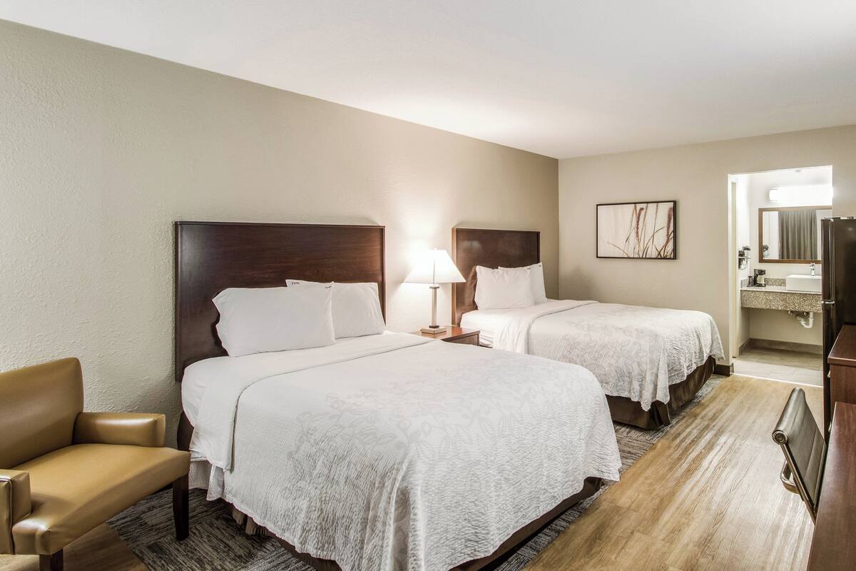 A hotel room with two beds, a chair, a desk, and a bathroom visible through an open door.
