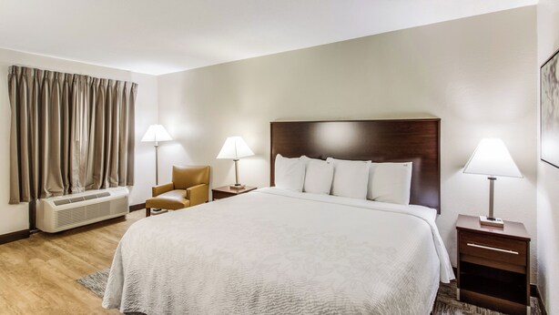 A hotel room with a large bed, two bedside tables with lamps, a chair, an air conditioning unit, and a window with curtains. - Red Roof Inn PLUS+ Dallas – Addison (Addison)