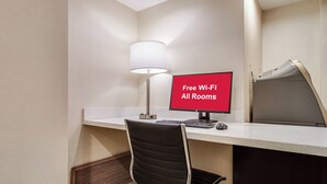 A hotel room with a desk, a chair, a computer, a lamp, and a sign indicating free Wi-Fi availability. - Red Roof Inn PLUS+ Dallas – Addison (Addison)