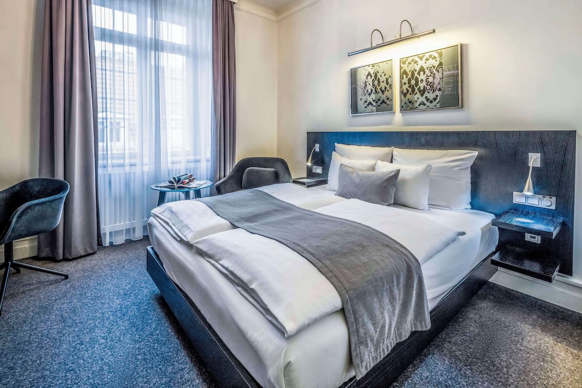 A hotel room with a large bed, a desk, a chair, a nightstand, and a window with curtains.
