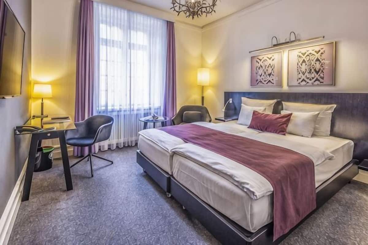 A hotel room with a bed, two bedside tables, a desk, a chair, and a window with curtains.