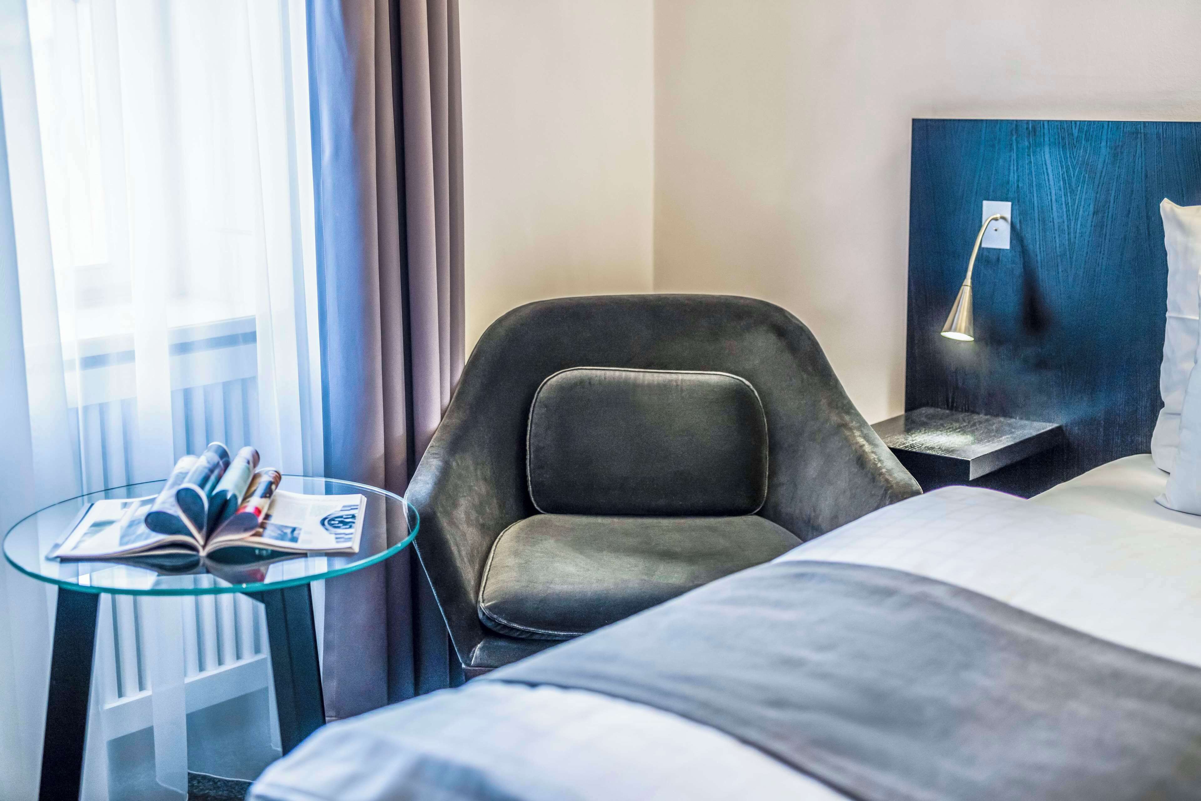 A hotel room with a bed, a glass table with magazines, a leather chair, a nightstand with a lamp, and a view of the outdoors.