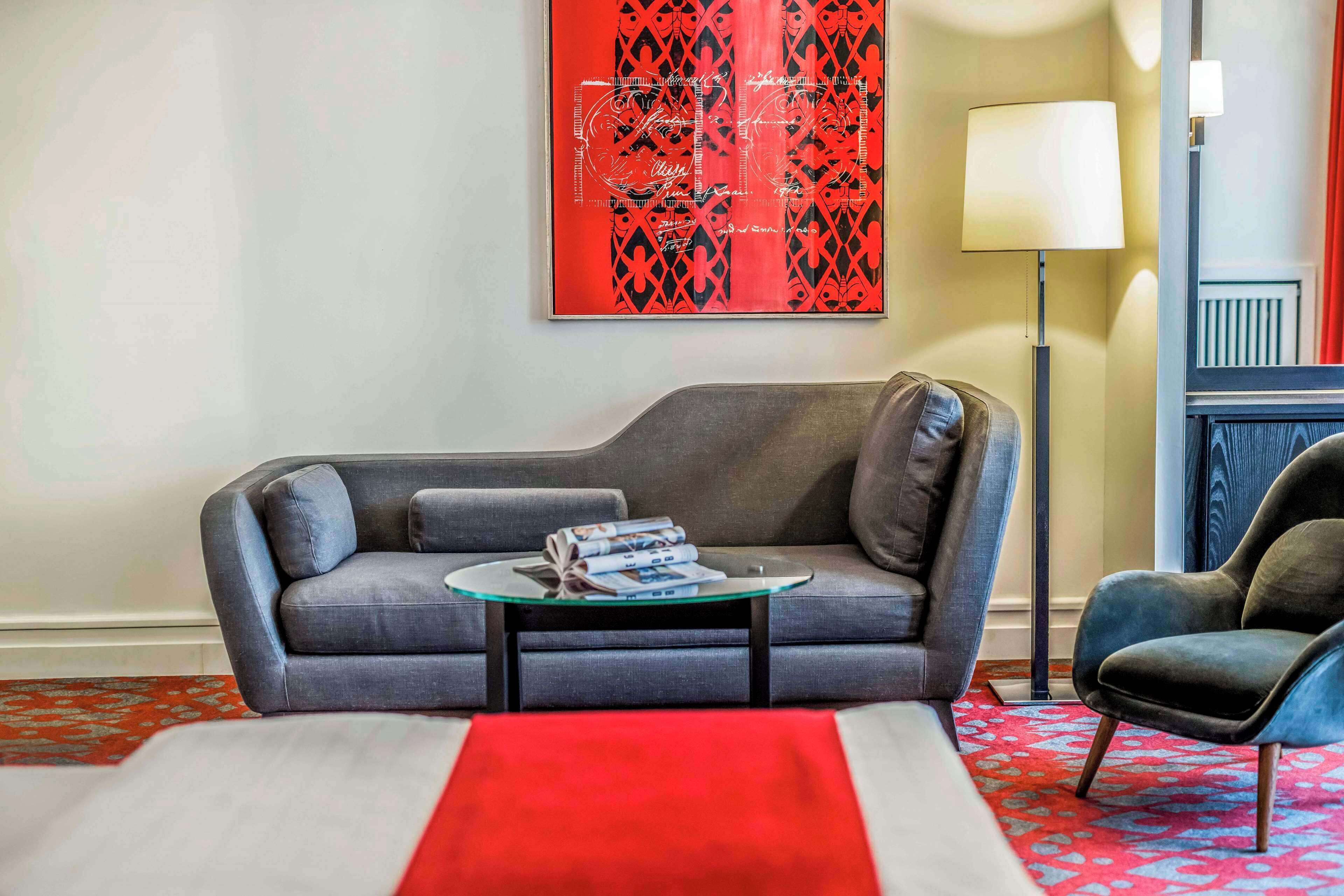 Ein modernes Wohnzimmer mit einem grauen Sofa, einem Glastisch mit Zeitschriften, einer roten Tagesdecke und einem roten Teppich mit Muster.