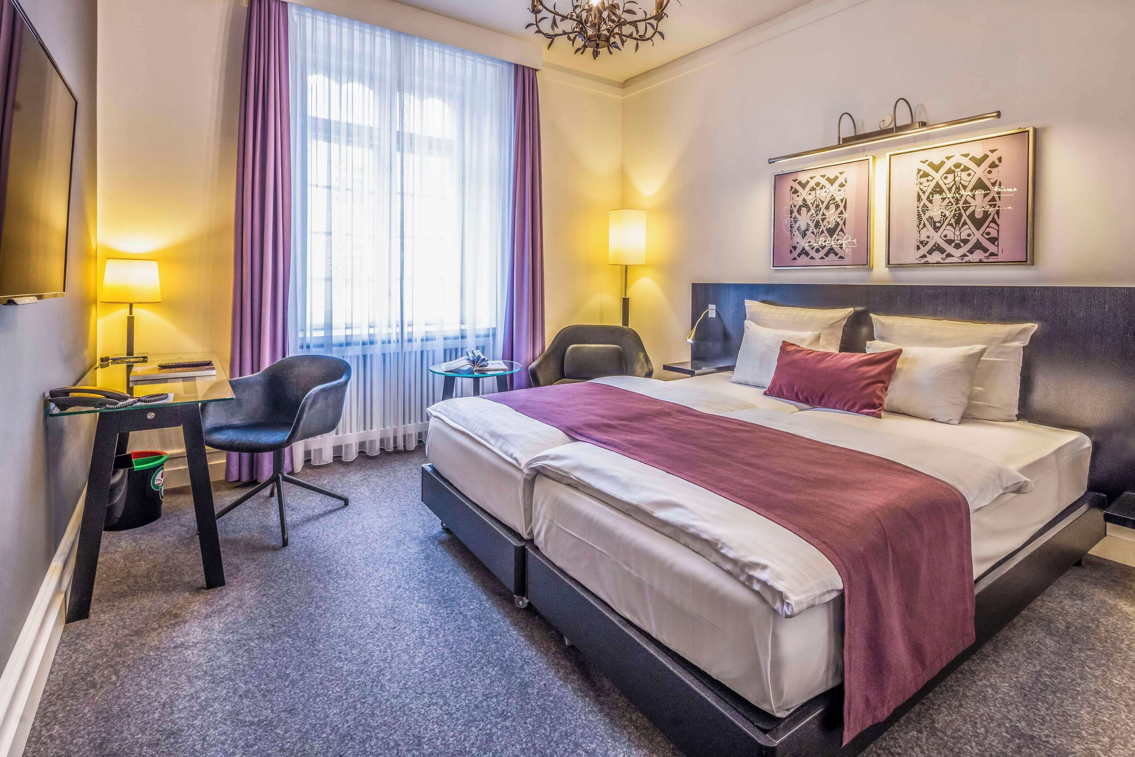 A hotel room with a bed, two chairs, a desk, and a window with curtains.