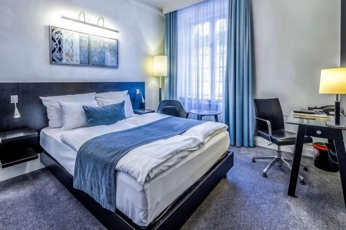 A hotel room with a bed, desk, chair, lamp, and artwork on the wall.