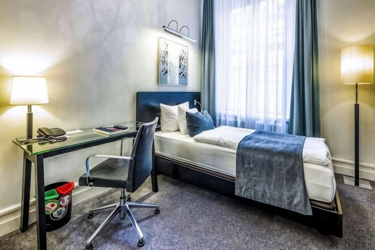 A hotel room with a bed, desk, chair, and a lamp.