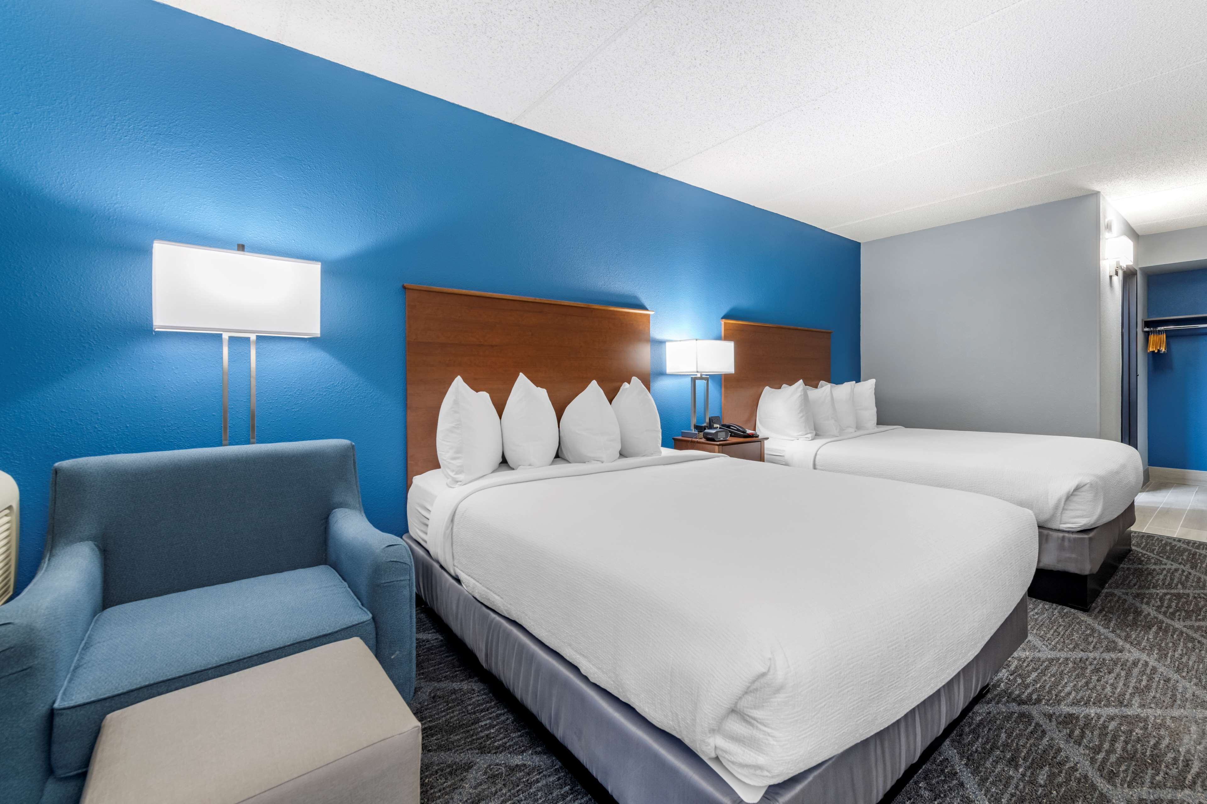 Quarto de hotel com duas camas, uma poltrona azul, uma mesinha e uma luminária.